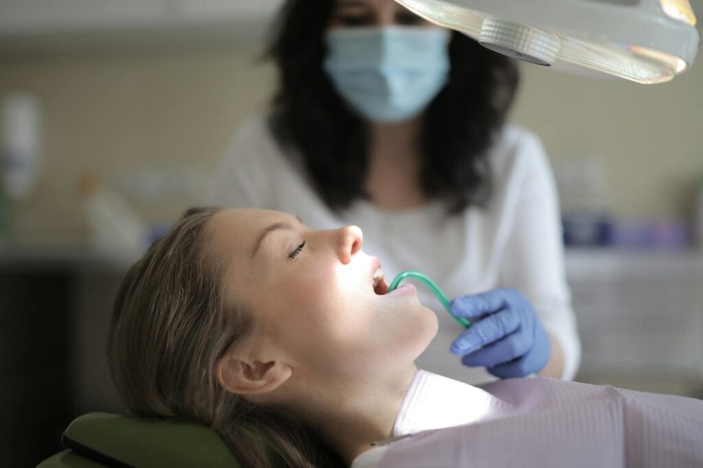 Child patient reclined in dental chair with mouth open, dentist wearing mask and gloves overhead lamp above.