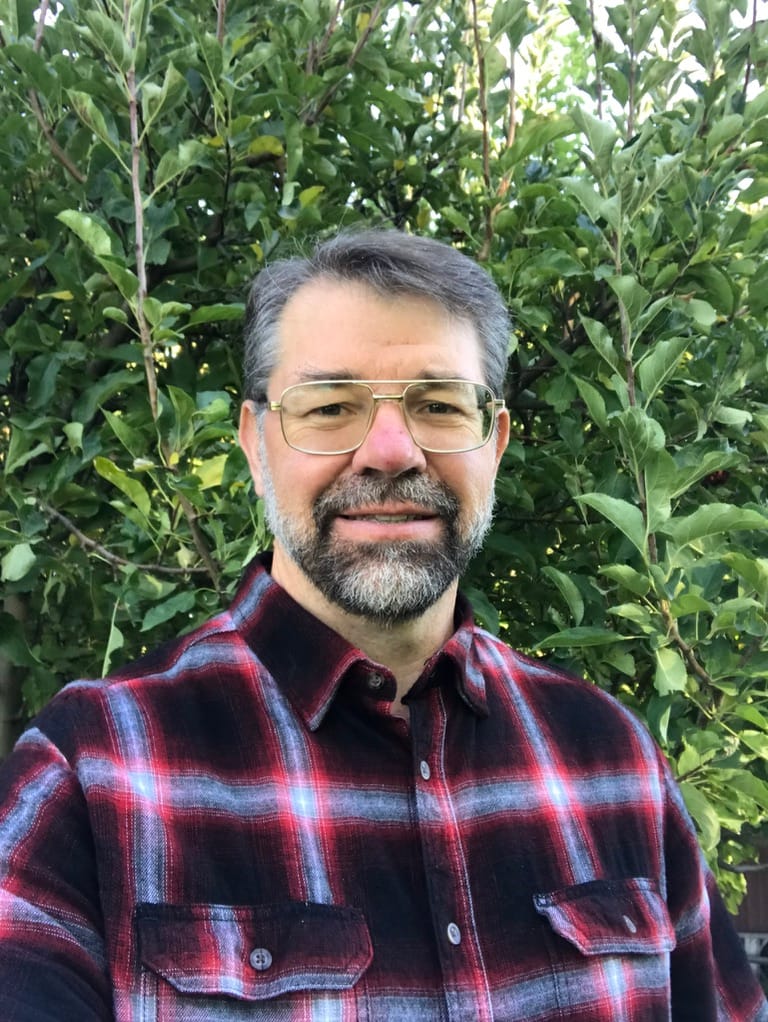 Portrait of a middle-aged man with gray beard and glasses, wearing a red plaid flannel shirt, outdoors with leafy bushes in the background.