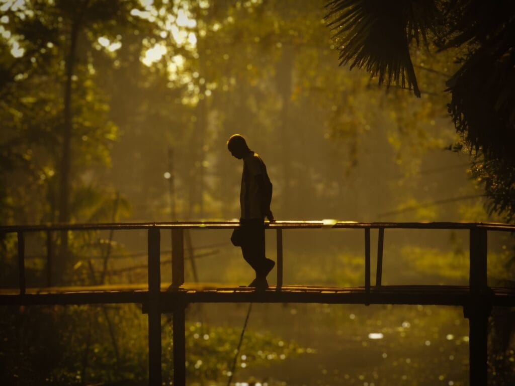 Silhouette of a man with his head lowered, walking across a small wooden footbridge in golden morning light